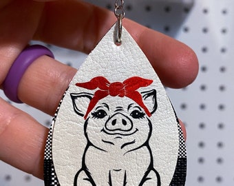 May include: White and black checkered leather earrings with a white pig wearing a red bandana on top. The pig is sitting down with its legs crossed.