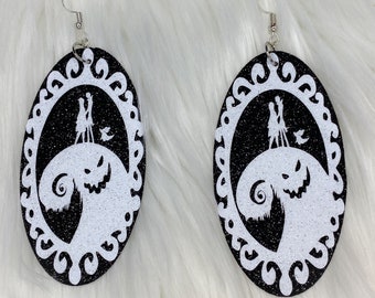 May include: A pair of black and white glitter earrings featuring a silhouette of Jack Skellington and Sally from the Nightmare Before Christmas movie. The earrings are oval shaped and have a white glitter frame.