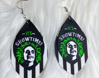 May include: A pair of black and white striped teardrop-shaped earrings with a green and black image of a character with the text "It's Showtime!" on the front.
