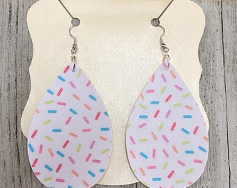 May include: A pair of white teardrop-shaped earrings with a colorful sprinkle pattern. The earrings are made of leather and have silver hooks.