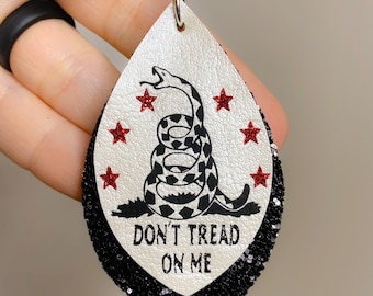 May include: A pair of dangle earrings with a white leather teardrop shape. The teardrop is decorated with a black and white snake with the words "Don't Tread On Me" and five red stars. The bottom half of the teardrop is black glitter.