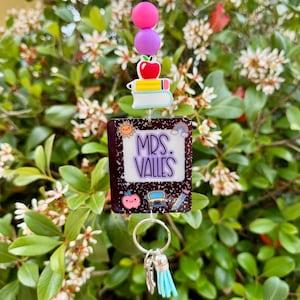 May include: A teacher-themed keychain with a dark purple notebook design that reads "MRS. VALLES". The keychain features an apple, books, a school bus, and a pencil charm. It includes a silver keyring and a light blue tassel.