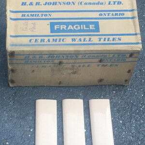 May include: A cardboard box labeled "H. & R. JOHNSON (Canada) LTD." with text including "FRAGILE" and "CERAMIC WALL TILES." Three rectangular, pale peach ceramic tiles are below the box. The box is aged and worn.