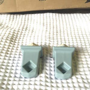 May include: Two light blue ceramic wall-mounted soap dishes. Each has a rectangular shape with a diamond-shaped opening and a small, angled top. They are set against a white, textured background.