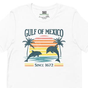May include: White t-shirt featuring a retro design with the text "Gulf of Mexico" above a sunset graphic with palm trees and dolphins. The text "Since 1672" is below the graphic. The shirt has a crew neck and short sleeves.