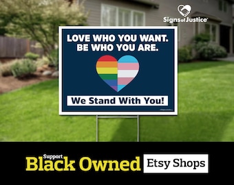 May include: A blue yard sign with the text "LOVE WHO YOU WANT. BE WHO YOU ARE." and a rainbow and transgender pride heart. The text "We Stand With You!" is below the heart. The sign is on a green lawn.
