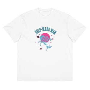May include: A white short-sleeve t-shirt featuring a central graphic of a light blue rearing horse against a solid pink circle. The text "SELF-MADE MAN" is arched above in light blue, surrounded by several dark purple stars on the shirt front.