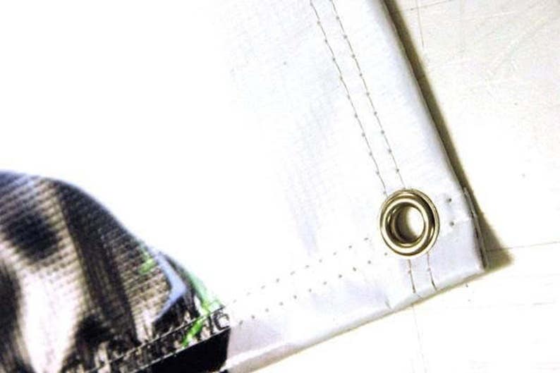 May include: Close-up of a white fabric banner with a metal grommet. The banner has a stitched edge and a subtle pattern of small dots.