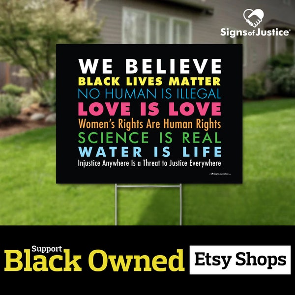 May include: Black yard sign with the text "We Believe Black Lives Matter No Human Is Illegal Love Is Love Women's Rights Are Human Rights Science Is Real Water Is Life" in colorful text on a black background. The sign is in front of a green lawn and a house.