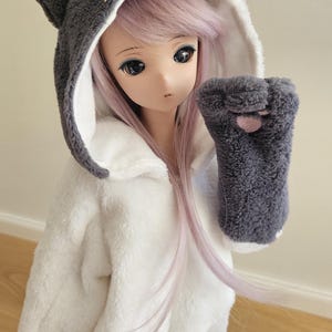 Könnte beinhalten: Eine Puppe in einem weiß-grauen Katzen-Overall. Das Outfit mit Kapuze hat graue Ohren, ein weißes Gesicht und Schnurrhaare. Die Puppe hat lange, hellviolette Haare und trägt einen grauen pfotenförmigen Fäustling.