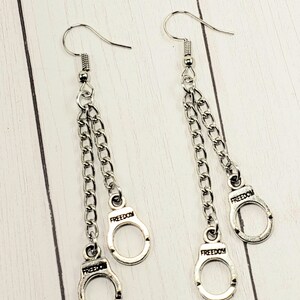 May include: Silver dangle earrings with two chains and three silver handcuff charms. Each handcuff charm has the word "FREEDOM" engraved on it.