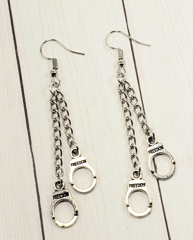 May include: Silver dangle earrings with two chains and three silver handcuff charms. Each handcuff charm has the word "FREEDOM" engraved on it.