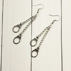 May include: A pair of silver dangle earrings with two handcuff charms on each earring. The handcuffs are connected to the earring by a silver chain.