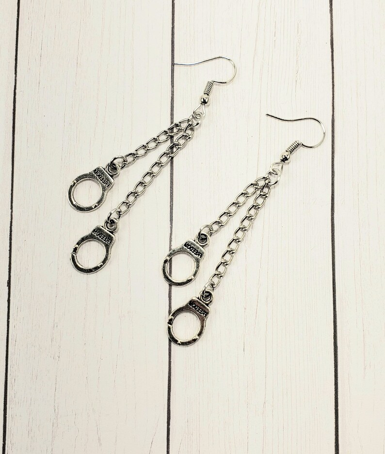 May include: A pair of silver dangle earrings with two handcuff charms on each earring. The handcuffs are connected to the earring by a silver chain.
