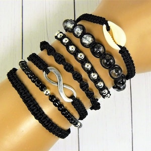 Puede incluir: Un conjunto de seis pulseras negras con diferentes encantos, incluyendo un símbolo de infinito, cuentas plateadas y un encanto de concha.