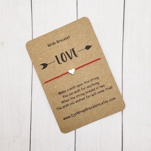 May include: Red string bracelet with a white heart charm and the word "LOVE" printed on a brown card. The card also includes instructions for making a wish on the string.