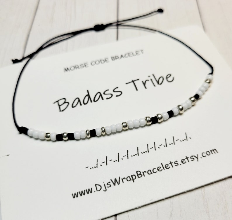 May include: A black string bracelet with white and black beads that spells out "Badass Tribe" in Morse code. The bracelet is on a white card with the text "Morse Code Bracelet" and "www.DJsWrapBracelets.etsy.com".