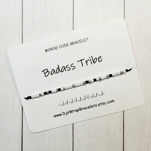 May include: A white card with black text that reads "Morse Code Bracelet" and "Badass Tribe". A black string bracelet with white and silver beads is displayed on the card. The text "www.DJSWrapBracelets.etsy.com" is printed on the card.