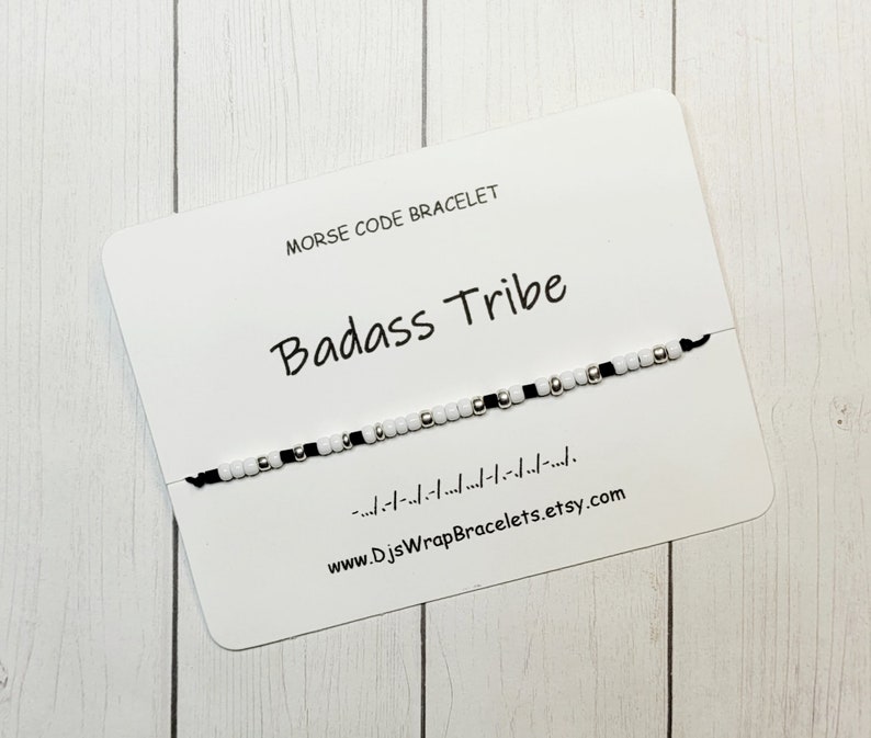 May include: A white card with black text that reads "Morse Code Bracelet" and "Badass Tribe". A black string bracelet with white and silver beads is displayed on the card. The text "www.DJSWrapBracelets.etsy.com" is printed on the card.
