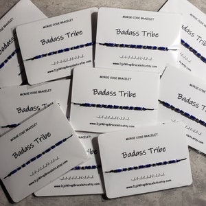 May include: Multiple white cards with the text "Morse Code Bracelet Badass Tribe" and a blue and silver beaded bracelet on each card. The text "www.DjsWrapBracelets.etsy.com" is printed on each card.
