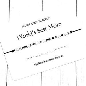 Könnte beinhalten: Ein weißes und schwarzes Perlenarmband mit der Aufschrift "World's Best Mom" in schwarzer Schrift. Das Armband befindet sich auf einer weißen Karte mit dem Text "Morse Code Bracelet" und einer Website-Adresse.