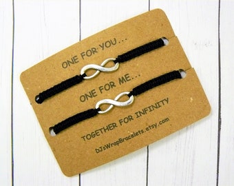May include: Two black cord bracelets with silver infinity charms are displayed on a brown card. The card reads "ONE FOR YOU... ONE FOR ME... TOGETHER FOR INFINITY" with a website address.