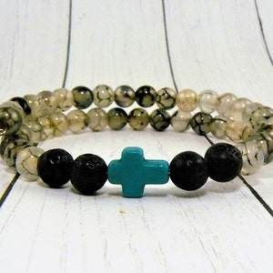 Puede incluir: Una pulsera de cuentas con dos filas de cuentas grises y blancas estampadas. La pulsera también incluye cuentas de lava negras y una cuenta en forma de cruz turquesa. Las cuentas están ensartadas en un cordón elástico negro.