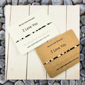 Pu&ograve; includere: Due braccialetti con perline bianche e nere che compongono la frase "Ti amo" in codice Morse su uno sfondo bianco e marrone. I braccialetti sono esposti su cartoncini con il testo "Braccialetto in codice Morse" e "Ti amo".