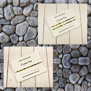 Pu&ograve; includere: Due braccialetti in corda nera con perline gialle che compongono "Ti amo" in codice Morse su un cartoncino bianco con il testo "Braccialetto in codice Morse Ti amo www.DjsWrapBracelets.etsy.com"