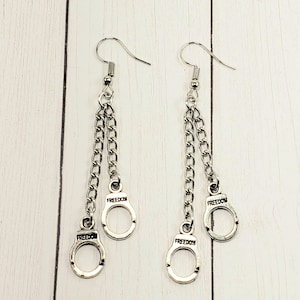 May include: Silver dangle earrings with two chains and three silver handcuff charms. Each charm has the word "FREEDOM" engraved on it.