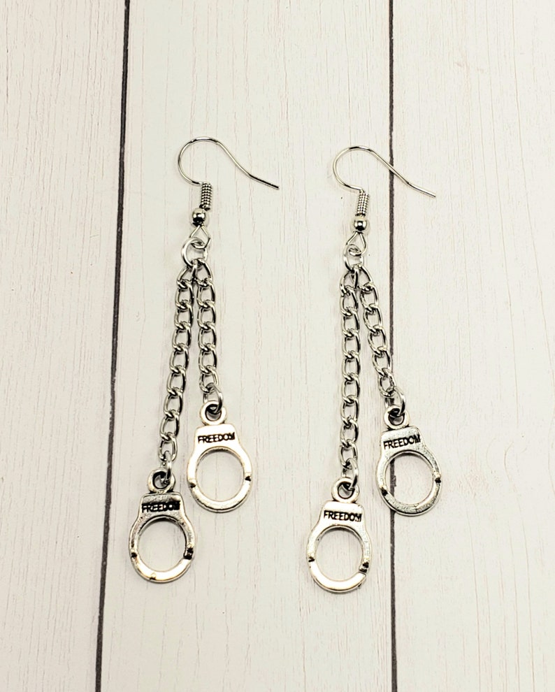 May include: Silver dangle earrings with two chains and three silver handcuff charms. Each charm has the word "FREEDOM" engraved on it.