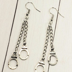 May include: Silver dangle earrings with two chains and a handcuff charm on each side. The handcuff charms have the word "FREEDOM" engraved on them.