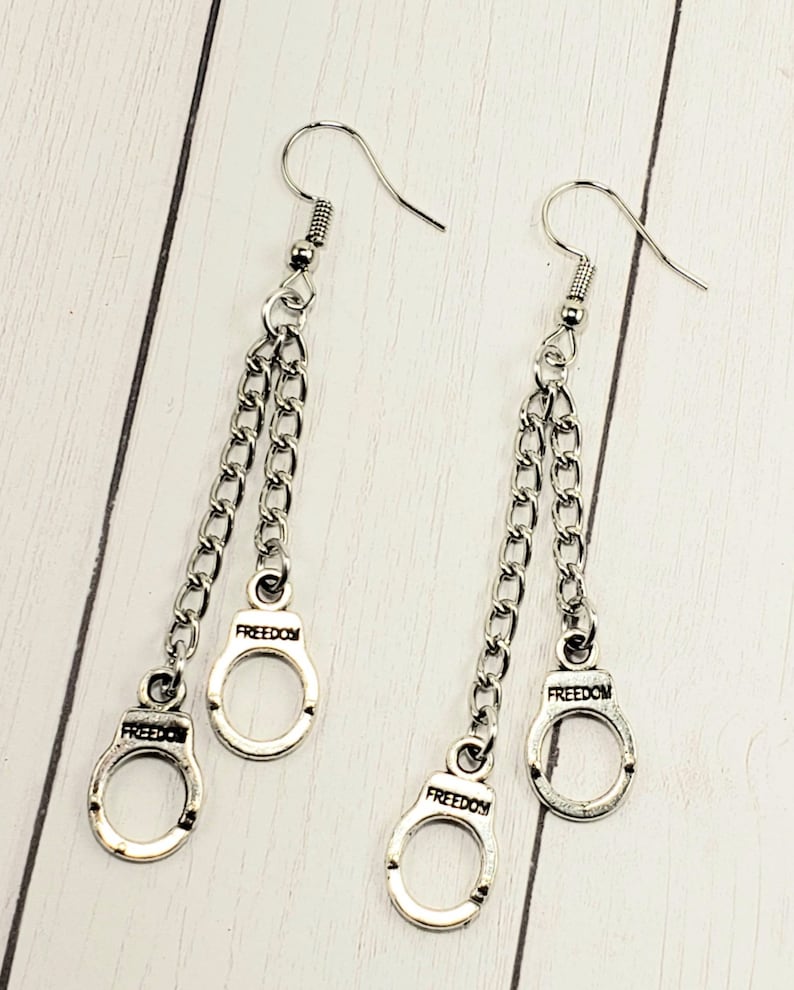 May include: Silver dangle earrings with two chains and a handcuff charm on each side. The handcuff charms have the word "FREEDOM" engraved on them.