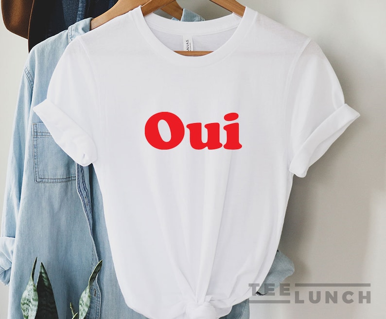 K&ouml;nnte beinhalten: Wei&szlig;es T-Shirt mit dem Wort "Oui" in fetten roten Buchstaben. Die &Auml;rmel sind hochgekrempelt und das T-Shirt ist an der Taille geknotet. Das T-Shirt h&auml;ngt an einem Kleiderst&auml;nder mit einem Jeanshemd.