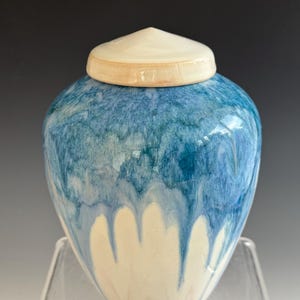 May include: A ceramic urn with a blue and white glaze. The urn has a rounded body and a conical lid. The blue glaze drips down the sides, creating a unique pattern. The lid is a creamy off-white color.
