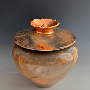 Peut inclure: Un pot en céramique fait à la main avec un couvercle. Le pot est de couleur marron chaud avec des accents orange et noirs. Le couvercle a un bord cuivré et un bord décoratif festonné. Le pot est une pièce de poterie unique.