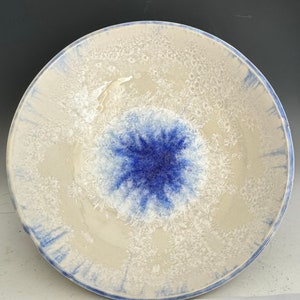 Op de afbeelding: Een witte keramische kom met een blauw en wit glazuur. De kom heeft een getextureerd oppervlak en een blauw centrum.