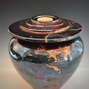May include: A dark, iridescent ceramic urn with a lid featuring concentric rings. The lid has a small, circular indentation holding a lit candle. Gold-colored accents adorn the urn's surface, adding a touch of elegance.