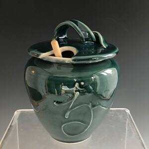 Puede incluir: Jarra de cerámica verde oscuro hecha a mano con tapa y asa. La jarra presenta un diseño decorativo en espiral y una pequeña cuchara de madera. La jarra está esmaltada y tiene un acabado brillante, ideal para artículos de cocina.