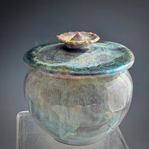 Op de afbeelding: Een handgemaakte keramische pot met een deksel. De pot heeft een ronde vorm met een brede opening en een deksel met een kleine, decoratieve knop. De pot heeft een glanzende, iriserende afwerking met tinten blauw, groen en paars.