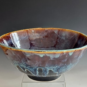 May include: A handmade ceramic bowl with a glossy, multi-toned glaze. The bowl features a dark brown interior with blue and teal accents, and a brown rim. The bowl sits on a clear surface against a dark background.