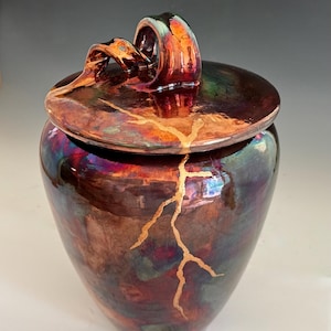 May include: A ceramic jar with a lid featuring a dark, iridescent, multi-colored glaze in shades of deep red, purple, and green. A prominent gold-colored crack repair line runs vertically across the lid and down the front of the jar body.