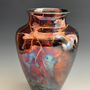 Puede incluir: Un jarrón de cerámica iridiscente hecho a mano con un exterior marrón oscuro y un acabado brillante. El jarrón presenta un diseño único con detalles dorados que resaltan las grietas, agregando un toque de elegancia a la pieza decorativa.