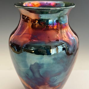 May include: A glass vase with a flared rim and a rounded body, featuring a highly reflective, iridescent finish that shifts between deep copper, bronze, and metallic blue tones, creating a swirling, multi-colored surface effect throughout.