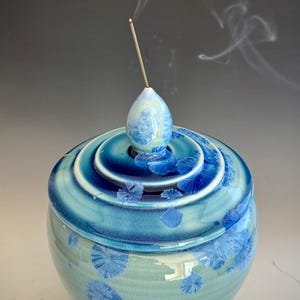 May include: A blue ceramic incense burner with a lid and a decorative finial. The burner has a crystalline pattern and is emitting smoke from a lit incense stick. The color palette includes shades of blue and white.