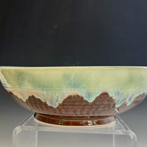 May include: A ceramic bowl featuring a two-tone glaze design. The upper portion displays a smooth, light green and pale yellow drip pattern, while the lower section has a textured, dark reddish-brown finish with a visible circular foot base.