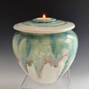 May include: A white ceramic urn with a teal and green glaze. The urn has a lid with a small opening for a tea light candle. The candle is lit.