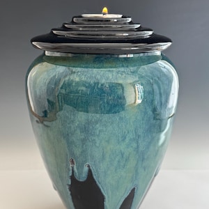 Op de afbeelding: Een keramische pot met een glanzend blauwgroen glazuur dat over een effen zwarte basis druipt. De houder heeft een getrapt zwart deksel met een klein, brandend theelichtje dat in het midden van het bovenoppervlak is geplaatst.
