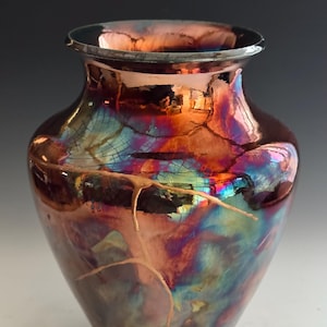 Könnte beinhalten: Eine dekorative Vase mit einzigartigem schillerndem Finish. Die Vase hat eine dunkelbraune Grundfarbe mit leuchtenden Farbtönen von Orange, Blau und Lila. Die Vase hat einen metallischen Rand und ein Craquelé-Design mit goldenen Akzenten.