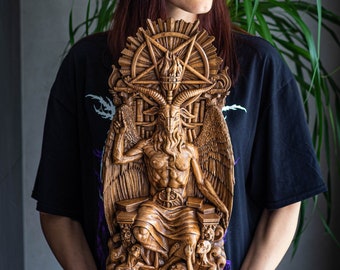 May include: A woman holds a wooden carving of a goat-headed figure with wings and a pentagram. The figure is seated on a throne and surrounded by smaller figures. The carving is detailed and intricate.